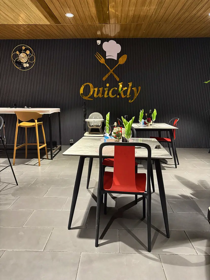 Le logo Quickly en lettres dorées sur un mur sombre, avec toque et couverts, au-dessus d’une table et de sa chaise rouge.